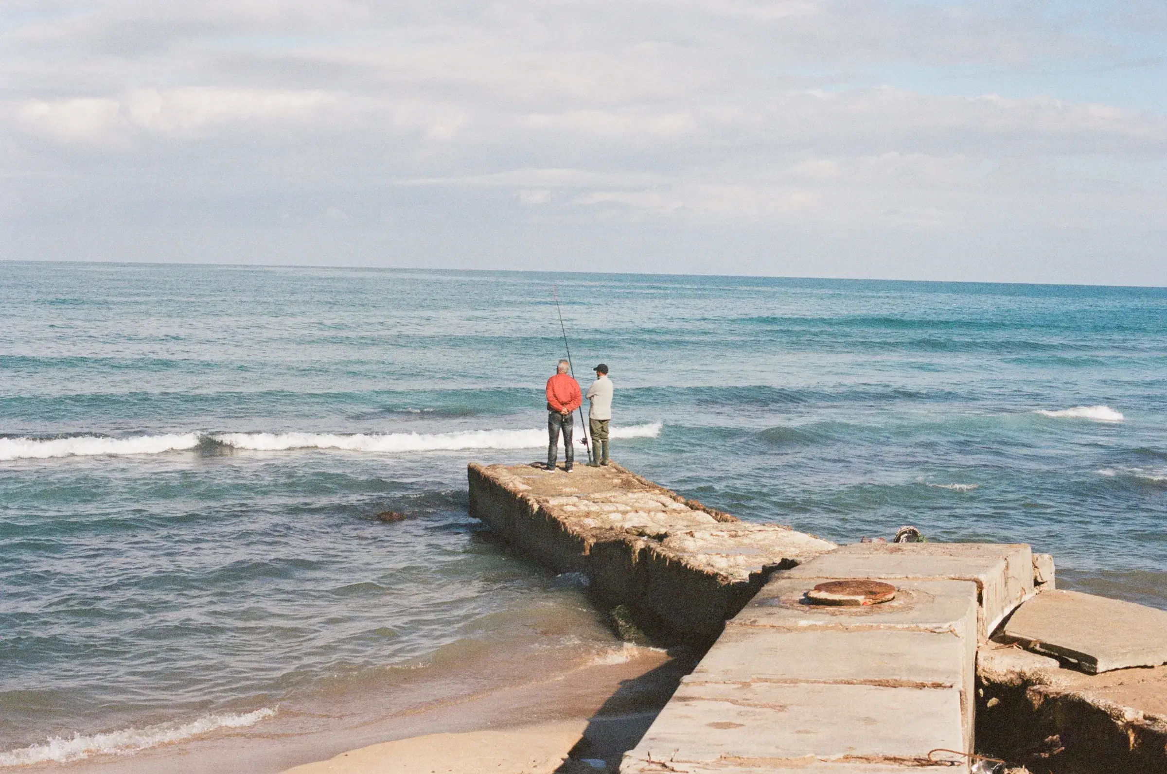 two figures at the end of a concrete pier, turquoise sea breaking around them
